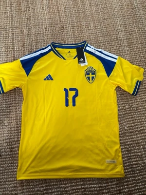 Svensk landslagströja Adidas gul Gyökeres M - Gul svensk landslagströja från Adidas med blå detaljer och nummer 17 på bröstet. Tröjan har blått och vitt mönster på axlarna, broderad Sverige-logga och Adidas-logga. Tillverkad i lätt och ventilerande material, perfekt för fotboll eller träning.Storlek M