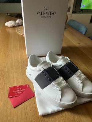 Valentino Garavani vita sneakers - Stilrena vita sneakers från Valentino Garavani med bred svart läderdetalj över vristen. Klassisk låg modell med vita snören och diskret logga på sulan. Tillverkade i slätt läder med gummisula och nitar på hälen för extra edge.