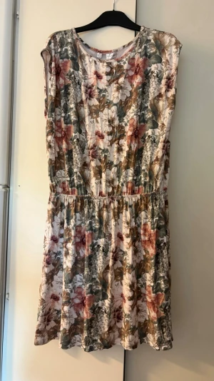 Blommig kortklänning i mjuk bomull - Supersöt kortklänning med blommigt mönster i beige, grön, rosa och vit. Klänningen har rund hals, resår i midjan och är ärmlös. Perfekt för varma dagar och enkel att styla med sneakers eller sandaler.