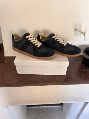 Maison Margiela - Snygga marinblå sneakers med ovandel i mocka och släta partier i läder. Skorna har vita snören och en klassisk gummisula i brunt. Insidan är ljusbeige vilket ger en fräsch kontrast. Perfekta för dig som gillar stilrena och tidlösa sneakers.