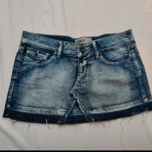 Blå jeanskjol med mörk kant - Cool kort jeanskjol i blå denim med slitna detaljer och rå, fransig kant. Kjolen har klassiska fickor fram och bak samt bälteshällor. Perfekt för en avslappnad och trendig look.