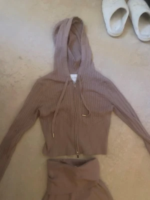 Beige ribbat loungewear set med huva - Mysigt loungewear set i beige med ribbad zip-hoodie och matchande vida byxor med hög midja. Hoodien har dragsko och guldiga detaljer på snörena. Perfekt för chill dagar hemma eller när du vill vara bekväm men ändå snygg. Använd 1 gång 
