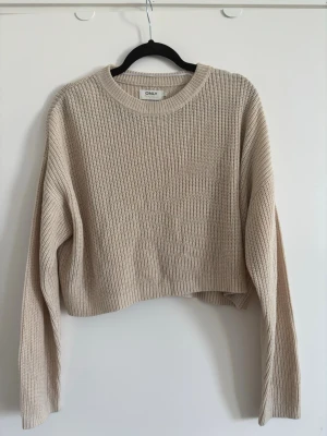 Beige cropped stickad tröja ONLY - Trendig beige stickad tröja från ONLY med croppad passform och ribbstickning. Tröjan har rund halsringning och långa ärmar, perfekt för lager på lager och en avslappnad look. Storlek L men passar på mig som har S.