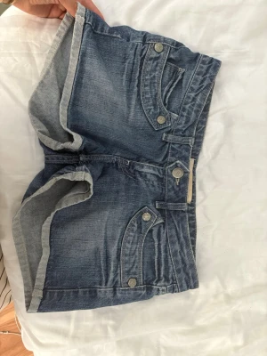 Blå lågmidjade jeansshorts med knappar - Snygga blå jeansshorts med uppvikta benslut och dekorativa knappar på fickorna. Klassisk femficksmodell med normal passform och midwaist. Perfekta för varma dagar och enkel att matcha med olika toppar. Står att de är i storlek M men är förmodligen i S då de är väldigt små. 