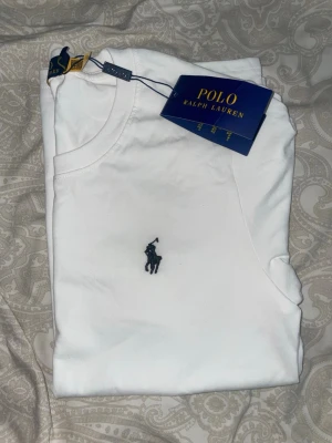 Vit t-shirt från Polo Ralph Lauren - Klassisk vit t-shirt från Polo Ralph Lauren med rund halsringning och ikonisk broderad logga på bröstet. T-shirten är i mjuk bomull och har en stilren, tidlös design som passar till allt.