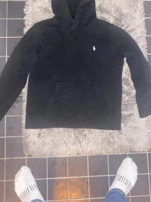 Svart hoodie från Polo Ralph Lauren - Klassisk svart hoodie från Polo Ralph Lauren med broderad vit logga på bröstet. Hoodien har en stor känguruficka framtill och en skön huva. Tillverkad i mjukt bomullsmaterial som känns riktigt bekvämt. Perfekt för en avslappnad och stilren look.