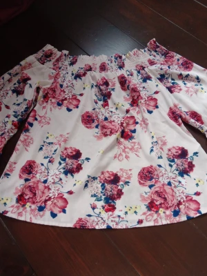 Offshoulder blommig blus i vitt och rosa - Supersöt offshoulder blus med vita och rosa toner, täckt av stora blommiga mönster i rosa, rött och blått. Blusen har resår upptill för en snygg passform och långa ärmar. Perfekt för dig som gillar romantisk och trendig stil.