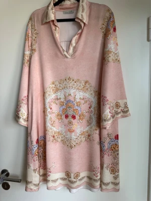 Rosa bohemisk klänning med mönster - Bohemisk klänning i ljusrosa med blommigt och ornamenterat mönster i beige, blått och rött. Klänningen har v-ringning, krage och vida ärmar.  Tillverkad i ett mjukt, lätt material viskos och silke  som känns lyxigt mot hudenPerfekt för sommarens alla tillfällen.