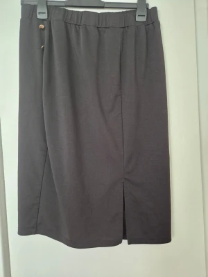Kjol - En enkel och stilren svart långkjol med resår i midjan och en diskret slits framtill. Kjolen har två små dekorativa knappar i midjan och är tillverkad i ett mjukt och bekvämt material. Perfekt för en avslappnad och trendig look.