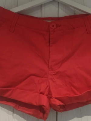 Röda shorts med uppvik - Snygga röda shorts med uppvikta benslut och bälteshällor. Klassisk knappstängning framtill och två dekorfickor bak. Perfekta för varma dagar och enkel att matcha med olika toppar.