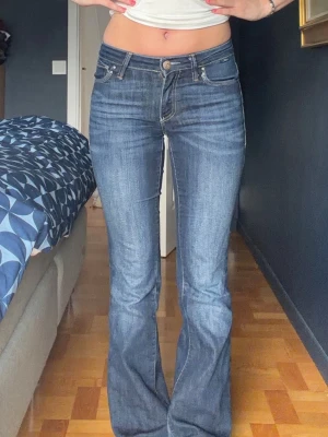 Blå bootcut jeans med låg midja - Säljer ett par blå bootcut jeans med låg midja och klassiska fem fickor. Jeansen har snygga kontrastsömmar och en lätt utsvängd passform nedtill. Perfekta för dig som gillar en retroinspirerad look. Materialet är denim i bomull.