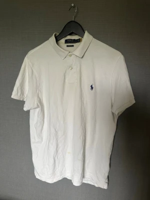 Vit piké från Polo Ralph Lauren - Klassisk vit pikétröja från Polo Ralph Lauren med korta ärmar och blå broderad logga på bröstet. Tröjan har krage och knappar framtill. Perfekt till en avslappnad men ändå stilren look.