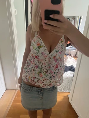 Blommig topp - Säljer en söt blommig linnetopp med v-ringning och tunna axelband. Toppen är vit med färgglada blommor och har spetsdetaljer samt broderade hålmönster. Perfekt för varma dagar och ger en romantisk vibe.