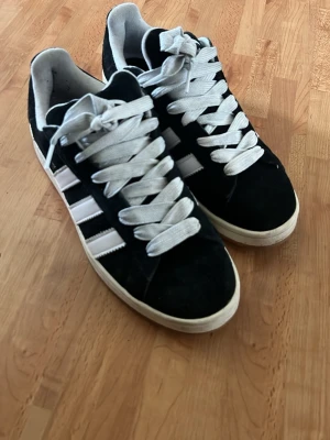 Svarta Adidas sneakers med vita ränder - Snygga svarta Adidas sneakers i mocka med klassiska vita ränder på sidorna och breda vita skosnören. Låg modell med vit sula och Adidas-logga på plösen. Perfekta för en streetwear-look och passar till det mesta.