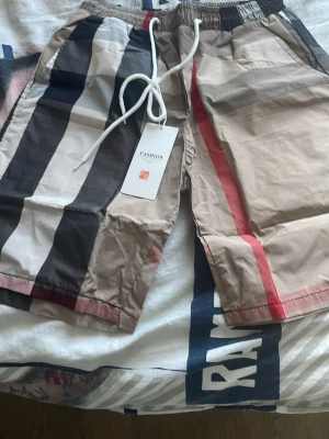 Randiga beige shorts med snörning - Säljer ett par snygga shorts med breda ränder i beige, svart, vitt och rött. De har elastisk midja med vit snörning och sidofickor. Perfekta för sommaren och riktigt sköna att bära till vardags. Stilen är inspirerad av klassiska streetwear-vibbar.