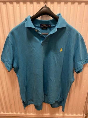 Blå pikétröja från Polo Ralph Lauren - Klassisk nästan helt ny blå pikétröja från Polo Ralph Lauren med korta ärmar och den ikoniska broderade loggan på bröstet. Tröjan har krage och knappar framtill, perfekt för en avslappnad men stilren look.