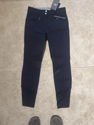 Marinblå ridbyxor Mountain Horse - Snygga marinblå ridbyxor från Mountain Horse, modell Diana Breeches. Byxorna har två dragkedjefickor fram, silikonprint på insidan av benen för extra grepp och dubbla knappar i midjan. Perfekta för ridning. 