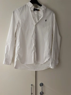 Vit skjorta från Ralph Lauren - Klassisk vit skjorta från Ralph Lauren med button-down krage och broderad logga på bröstet. Skjortan har lång ärm, bröstficka och knappar framtill. Perfekt till både jeans och kostymbyxor.