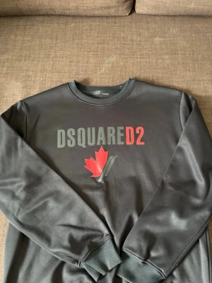 Svart sweatshirt från Dsquared2 - Cool svart sweatshirt från Dsquared2 med tryckt logga i grått och rött samt ett rött lönnlöv på bröstet. Tröjan har rund halsringning och ribbade muddar vid ärmslut och nederkant. Perfekt för dig som gillar streetwear och snygga detaljer.