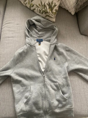 Polo Ralph Lauren zip hoddie  - Snygg grå hoodie från Polo Ralph Lauren med dragkedja framtill och klassisk blå logga på bröstet. Hoodien har huva och två stora fickor framtill. Tillverkad i mjuk bomullsblandning som känns skön mot huden.