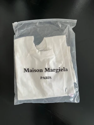 Vit Maison Margiela t-shirt - Snygg vit t-shirt från Maison Margiela med klassisk logga tryckt baktill och diskreta sömdetaljer framtill. T-shirten har rund hals och korta ärmar, tillverkad i mjuk bomull för en clean och stilren look.