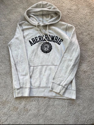 Grå hoodie från Abercrombie - Snygg ljusgrå hoodie från Abercrombie med svart broderad logga och emblem på bröstet. Klassisk känguruficka framtill och justerbar huva med dragsko. Perfekt för en avslappnad och trendig look. Mjuk bomullsblandning för extra komfort.