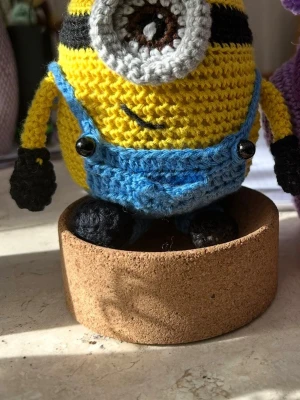Virkat Minion gosedjur - Handvirkat Minion gosedjur med gul kropp, blå hängselbyxor och svart detaljering. Figuren har ett stort grått öga med svart och vitt, samt svarta händer och fötter. Gjord av garn och dekorerad med knappar på byxorna. Perfekt som prydnad eller present.
