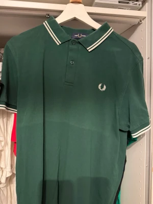 Fred Perry pike - Klassisk grön pikétröja från Fred Perry med vit logga broderad på bröstet. Tröjan har korta ärmar, krage med vita ränder och knäppning framtill. Perfekt för en stilren och sportig look.