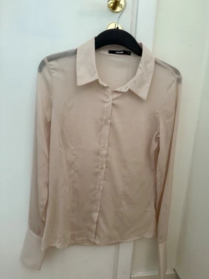 Beige skjorta från Bik Bok - Stilren beige skjorta från Bik Bok med klassisk krage och knappar framtill. Skjortan har lång ärm och är tillverkad i ett lätt, något glansigt material som ger en elegant känsla. Perfekt att styla lager på lager eller bära som den är. Den är genomskinlig 