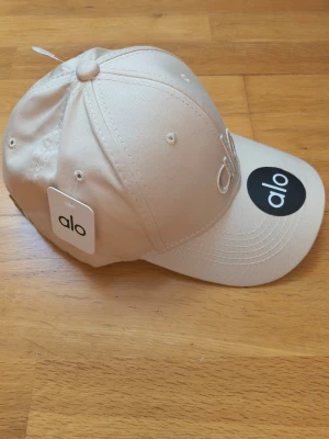 Beige keps från Alo med broderad logga - Snygg beige keps från Alo med broderad logga framtill. Klassisk böjd skärm och justerbar passform. Perfekt accessoar för en clean och sportig look. Kepsen är tillverkad i bomull och har ventilerande hål upptill.