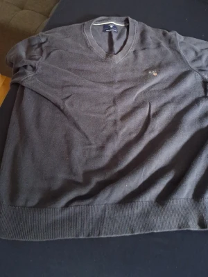Mörkgrå stickad tröja Gant - Snygg mörkgrå stickad tröja från Gant med V-ringning och diskret logga broderad på bröstet. Tröjan har ribbade muddar vid ärmslut och nederkant, perfekt för lager på lager. Klassisk och enkel design som passar till det mesta.