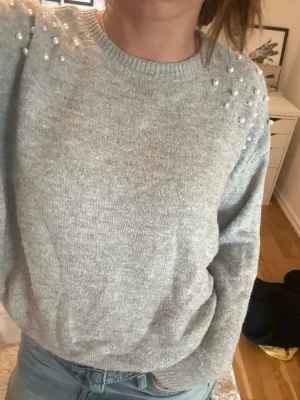 Ljusgrå stickad tröja med pärlor - Supersnygg ljusgrå stickad tröja med pärldetaljer på axlarna. Tröjan har rund halsringning och långa ärmar. Perfekt för dig som vill ha något både mysigt och trendigt. Materialet känns mjukt och skönt mot huden. Passar s + m