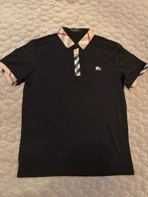 Svart Burberry pikétröja med rutig krage - Snygg svart pikétröja från Burberry med klassisk beige och röd rutig krage och knappslå. Ikonisk Burberry-logga broderad på bröstet. Kortärmad modell i mjukt material, perfekt för en stilren look.