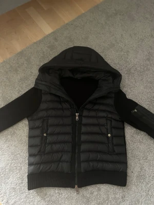 Moncler cardigan  - Snygg svart pufferjacka från Moncler med stickade ärmar och huva. Jackan har två fickor med knappar framtill och en dragkedja. Ärmarna är ribbstickade och har en ficka med Moncler-logga på ena ärmen. Perfekt för kalla vinterdagar.