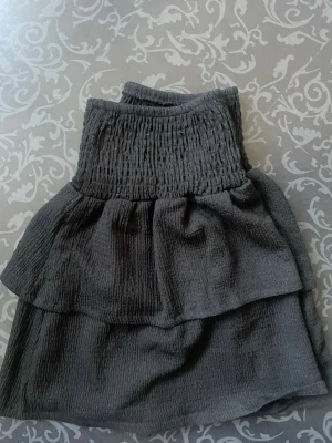 Svart volangkjol - Supersnygg svart kjol med bred smock i midjan och dubbla volanger. Kjolen är gjord i ett mjukt, strukturerat tyg som ger en avslappnad men trendig look. Perfekt att matcha med en crop top eller stickad tröja.
