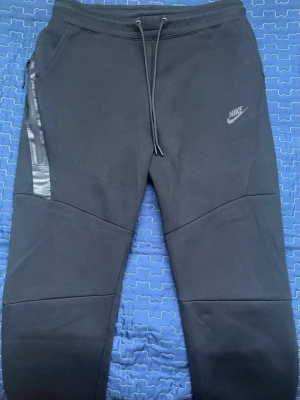 Svarta Nike Tech Fleece mjukisbyxor - Nike Tech Fleece mjukisbyxor i svart med smal passform och dragsko i midjan. Byxorna har en snygg detalj med dragkedja på sidan och diskret Nike-logga på låret. Tillverkade i mjukt och stretchigt material som ger en skön känsla hela dagen.