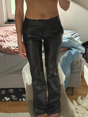 Svarta bootcut byxor i skinnimitation - Säljer ett par svarta byxor i skinnimitation med bootcut passform. Byxorna har hög midja och stängs med dragkedja och knapp framtill. Snygga att styla till crop tops eller oversized tröjor för en edgy look.