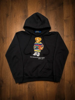 Svart hoodie Polo Bear Ralph Lauren - Svart hoodie från Ralph Lauren med den ikoniska Polo Bear-trycket på bröstet. Björnen har färgglada kläder och håller en basketboll. Hoodien har huva med dragsko och en stor magficka. Perfekt för dig som gillar streetwear och klassiska märken.