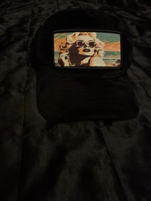 Svart keps med retrotryck - Säljer en svart keps med mesh på sidorna och ett stort retroinspirerat tygmärke framtill. Märket visar en kvinna med solglasögon i pop art-stil mot en strandbakgrund i färger som beige, orange och turkos. Perfekt för dig som gillar unik streetwear och coola detaljer.