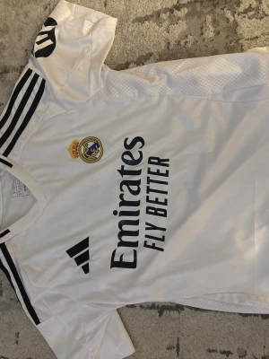 Real Madrid matchtröja Adidas vit - Vit Real Madrid matchtröja från Adidas med svarta detaljer och klubbmärke på bröstet. Tryck med 'Emirates Fly Better' och UEFA Foundation-märke på ärmen. Klassisk design med svarta ränder på axlarna. Perfekt för fotbollsfans! Tröjan passar som S