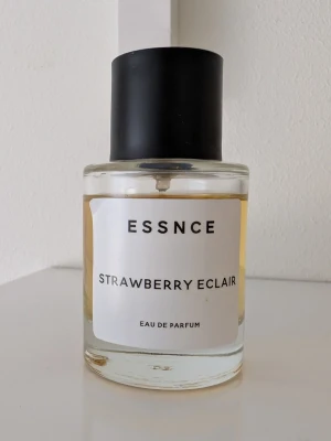 ESSNCE Strawberry Eclair parfym - Parfymen ESSNCE Strawberry Eclair kommer i en rund glasflaska med svart lock och vit etikett. Doften är inspirerad av jordgubb och bakverk, vilket ger en söt och fruktig känsla. Flaskan har en minimalistisk design och innehållet är ljusgul vätska.