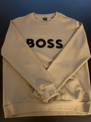 Beige sweatshirt från BOSS - Snygg beige sweatshirt från BOSS med svart logotyp tryckt framtill. Tröjan har rund halsringning, långa ärmar och ribbade muddar vid ärmslut och nederkant. Tillverkad i mjukt material som känns skönt mot huden. Perfekt för en avslappnad och trendig look.
