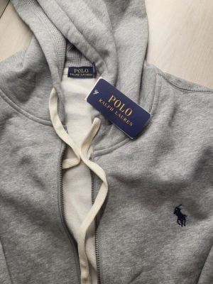 Grå hoodie från Polo Ralph Lauren - Snygg grå hoodie från Polo Ralph Lauren med dragkedja, vit dragsko och klassisk broderad logga på bröstet. Mjuk bomullsblandning och ribbade muddar. Perfekt för en chill och stilren look.