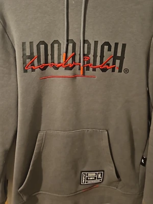 Grå HOODRICH hoodie med röd text - Snygg grå hoodie från HOODRICH med stor svart och röd logga på bröstet. Hoodien har huva med dragsko och en stor magficka. Detaljer i rött på snörena och en patch vid fickan. Perfekt för en streetwear-look. Säljer den för den har blivit för liten men inte mycket användning 