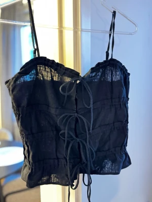 Svart linnetopp med snörning - Trendig svart linnetopp med tunna axelband och snygg snörning framtill. Toppen är tillverkad i ett lätt, luftigt material som passar perfekt till varma dagar. Enkel men ändå detaljerad design som ger en cool vibe. Endast testad
