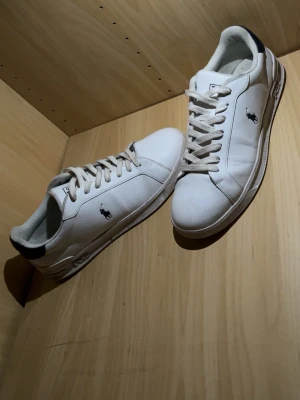 Vita Polo Ralph Lauren sneakers - Klassiska vita sneakers från Polo Ralph Lauren med lågt skaft, vita snören och mörkblå detaljer vid hälen. Ikoniska Polo-loggan på sidan och 'Polo' broderat bak. Tillverkade i slätt läder med gummisula. Perfekta för en clean och stilren look. Pris kan diskuteras.