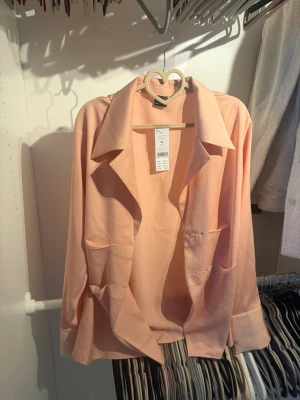 Peachfärgad satinblus Amra XL - Snygg peachfärgad skjorta från Amra i satinmaterial. Skjortan har en oversized passform, bred krage och stora fickor framtill. Perfekt för dig som gillar en avslappnad men trendig stil. Storlek XL.