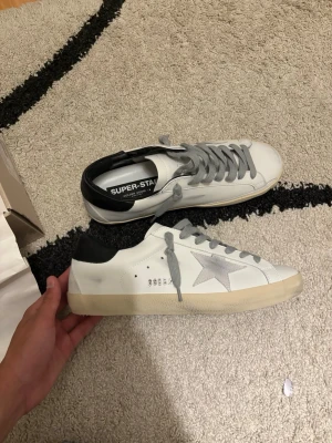Golden Goose Superstar vita sneakers - Golden Goose Superstar sneakers i vitt läder med grå stjärndetalj på sidan, grå skosnören och svart hälparti. Sulan är beige och har en vintage-look. Klassisk låg modell med perforerade detaljer och logga på sidan. Allt på bilderna kommer med, påse, box, golden goose passport. Skriv vid funderingar. Kan sänka pris vid snabb affär! 