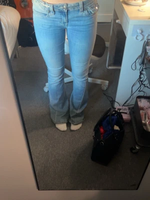 Ljusblå bootcut jeans med fickdetaljer - Säljer ett par ljusblå bootcut jeans med låg midja och snygga fickdetaljer bak med knapp. Jeansen har klassisk femficksmodell och är tillverkade i stretchigt denimtyg för skön passform. Perfekta för dig som gillar retrovibbar och en avslappnad look. Lite slitna i byxändorna men går lätt att ta bort. Där av lite billigare.