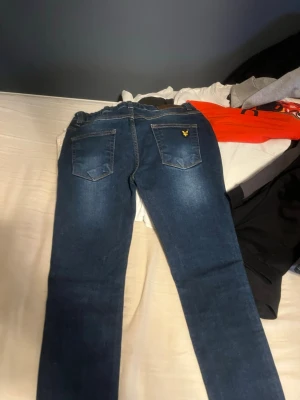 Mörkblå jeans från Lyle & Scott - Snygga mörkblå jeans från Lyle & Scott med klassisk femficksdesign och gul logga på ena bakfickan. Jeansen har raka ben och kontrastsömmar samt en liten röd detalj vid fickan fram. Perfekta för en avslappnad och trendig look.